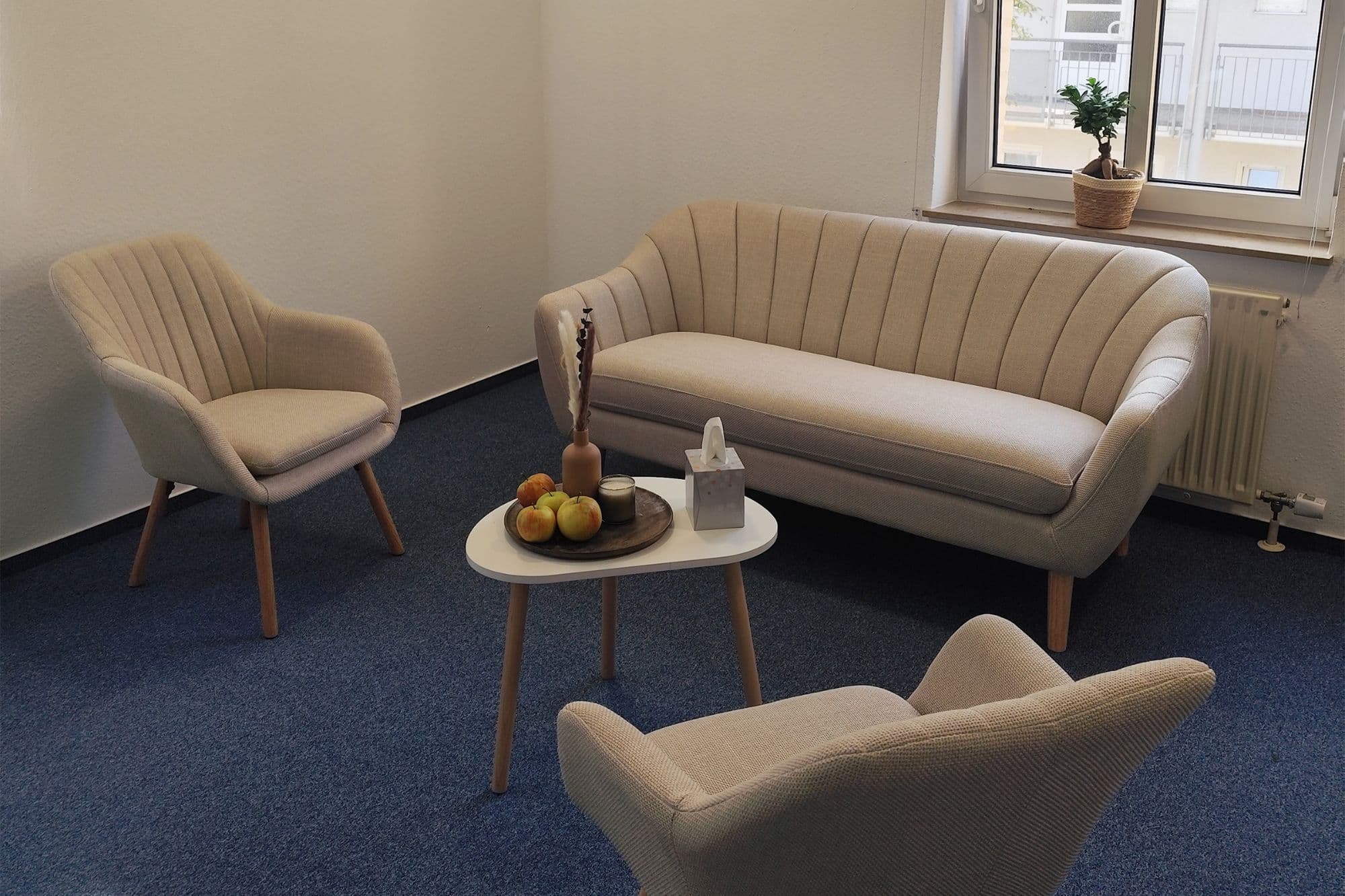 Praxisraum mit beigem Sofa, zwei Sesseln und einem kleinen Couchtisch vor einem Fenster